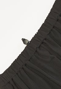 Svart stoff søm med elastisk rynkete design, en glatt tekstur, og en liten svart knappdetalj for justering.