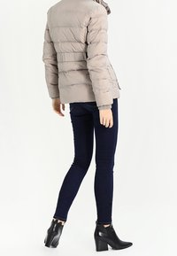 Mujer vistiendo una chaqueta acolchada beige, pantalones vaqueros ajustados azul oscuro y botas de tacón negro, de pie con la espalda vuelta.