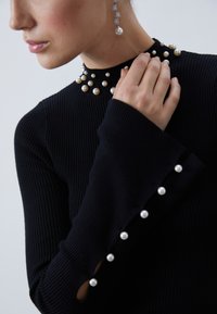Melns ribots džemperis ar apaļām pērļu rotājumiem pie apkakles un aprocēm, piedāvājot pieguļošu dizainu ar izplestiem piedurkņu galiem.