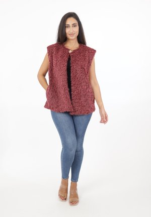 Faux fur vest i burgunderrød med krave, åben front og sidelommer. Båret over en sort top, parret med blå stramme jeans og gennemsigtige sandaler.