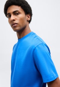 Νέος άνδρας με κοντά σγουρά μαλλιά που φοράει φωτεινό μπλε κοντομάνικο μπλουζάκι, κοιτάζει ελαφρώς πλάγια σε απλό φόντο.