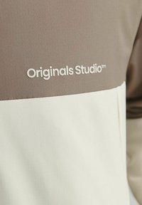 Ľahká bunda s hnedo-bielym farebným blokovaním, textúrovaná látka a logo "Originals Studio™" v bielej farbe.