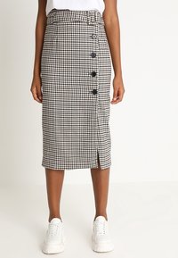 Falda midi de cuadros en blanco y negro con corte recto, que presenta una abertura frontal y cinco botones negros a lo largo del costado. Combinada con zapatillas blancas.