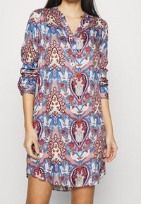 Nainen yllään pitkähihaista, napillista mekkoa, jossa on monivärinen kukka- ja geometrinen kuvio sinisen, punaisen, vaaleanpunaisen ja valkoisen sävyissä.