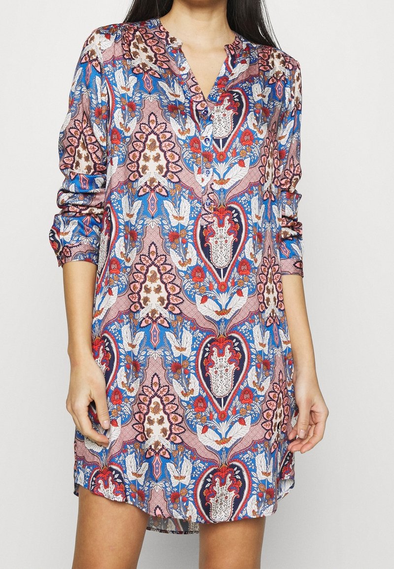 Nainen yllään pitkähihaista, napillista mekkoa, jossa on monivärinen kukka- ja geometrinen kuvio sinisen, punaisen, vaaleanpunaisen ja valkoisen sävyissä.