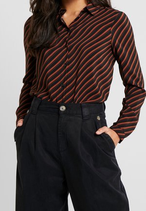 Femme portant une chemise à manches longues avec des rayures diagonales noires et rouille, rentrée dans un pantalon noir taille haute, les mains dans les poches.