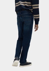 Jeans in denim blu scuro con taglio dritto, dotati di tasche posteriori e dettagli di cucitura sottili. Abbinati a sneaker nere e a una maglietta a righe.