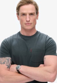 Grå T-shirt med korte ærmer, rund hals og en brystlomme. Har synlige sømme og et lille logo-mærke. Casual fit, bomuldsblanding.