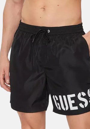 Costume da bagno nero con logo bianco "GUESS" sulla gamba sinistra, dotato di vita elasticizzata e coulisse, indossato da una persona senza maglietta.