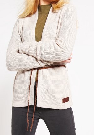 Kvinde iført beige strikket cardigan med brunt flettet bælte over olivengrøn top og mørkegrå jeans, med armene over kors.