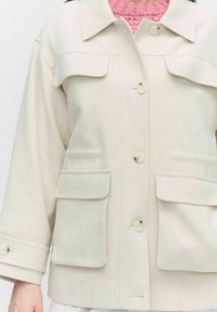 Ljusbeige jacka med knappstängning fram, två bröstfickor och vida ärmar. Texturerad tyg med subtila detaljer på knappar och ärmslut.