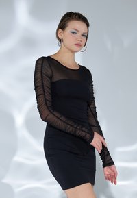 Musta mekko, jossa on tiukka muotoilu ja pitkät läpikuultavat hihat, joissa on rypytetty pinta, korostaen sulavaa kangasta vartalon alueella.