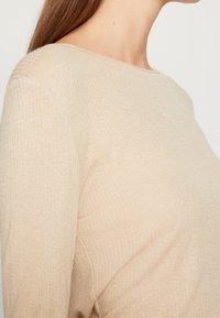 Femme aux longs cheveux bruns portant un haut beige côtelé à manches longues, montrée de l'épaule à la poitrine en profil latéral sur un fond uni.