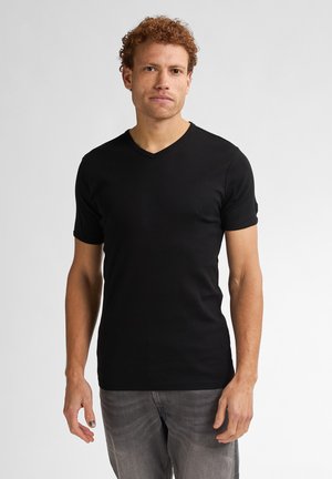 Man met krullend rood haar, die een eenvoudig zwart V-hals T-shirt en grijze spijkerbroek draagt, staand voor een effen witte achtergrond.