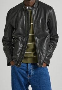 Giubbotto bomber in pelle nera con chiusura con zip, collo rotondo e tasche laterali, indossato sopra un maglione a righe verdi e jeans blu.