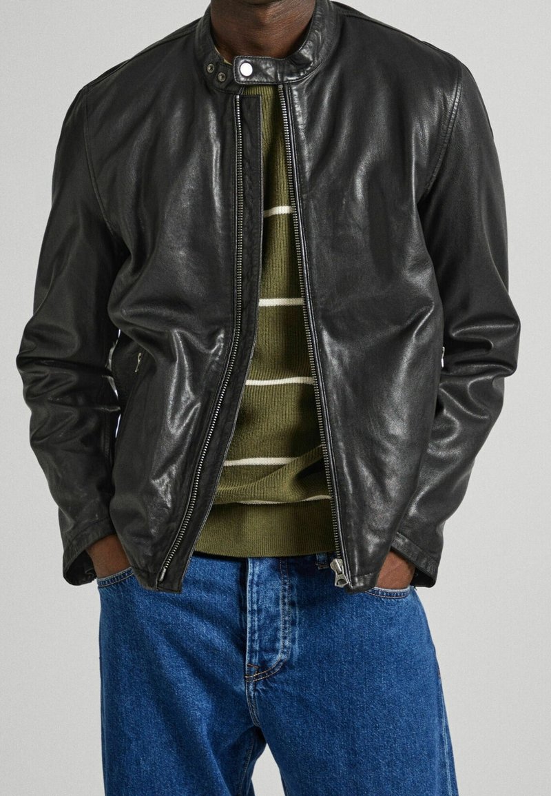 Giubbotto bomber in pelle nera con chiusura con zip, collo rotondo e tasche laterali, indossato sopra un maglione a righe verdi e jeans blu.