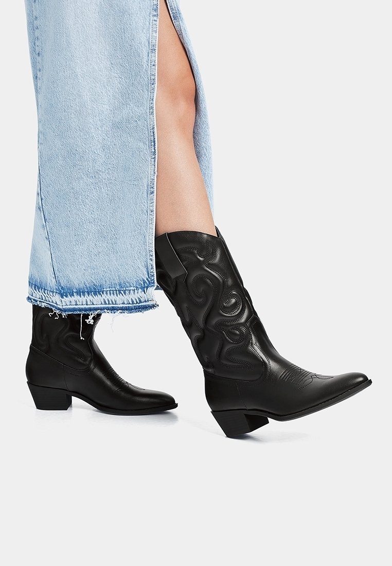 Bottes de cowboy en cuir noir avec un bout pointu, des détails texturés et un petit talon empilé, associées à une jupe en denim bleu clair.
