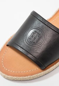 Sandale à enfiler en cuir noir avec semelle intérieure beige, coutures blanches, semelle en jute tressé et logo circulaire embossé sur la sangle.
