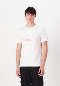 T-shirt alb din bumbac, cu text imprimat roz "LYLE & SCOTT ESTD 1874" curbat pe piept. Mâneci scurte și guler rotund.