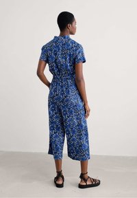 Zils ziedu raksts kombinezons no viegla auduma, ar īsām piedurknēm, savilktu jostasvietu un īsiem plašiem svārkiem. Iekļautas melnas sandales.