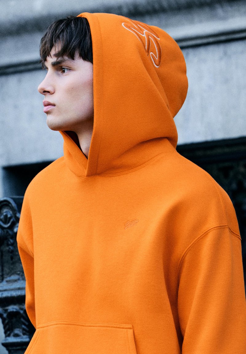 Nuori mies oranssissa hupullisessa svetarissa katsoo sivulle, rinnassa ja hupussa minimaalinen kirjailu, seisoo kivestä ja metallista rakenteiden lähellä.