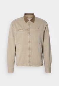 Chaqueta beige con un cuello de pana marrón, cierre frontal con cremallera y dos bolsillos laterales. Presenta un pequeño logo en el pecho y una tela texturizada.