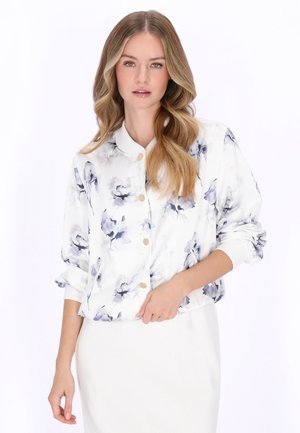 Frau mit langen blonden Haaren, die eine weiße, mit Blumen gemusterte, geknöpfte Jacke mit hochgekrempelten Ärmeln und einen weißen Rock trägt, vor einem weißen Hintergrund stehend.