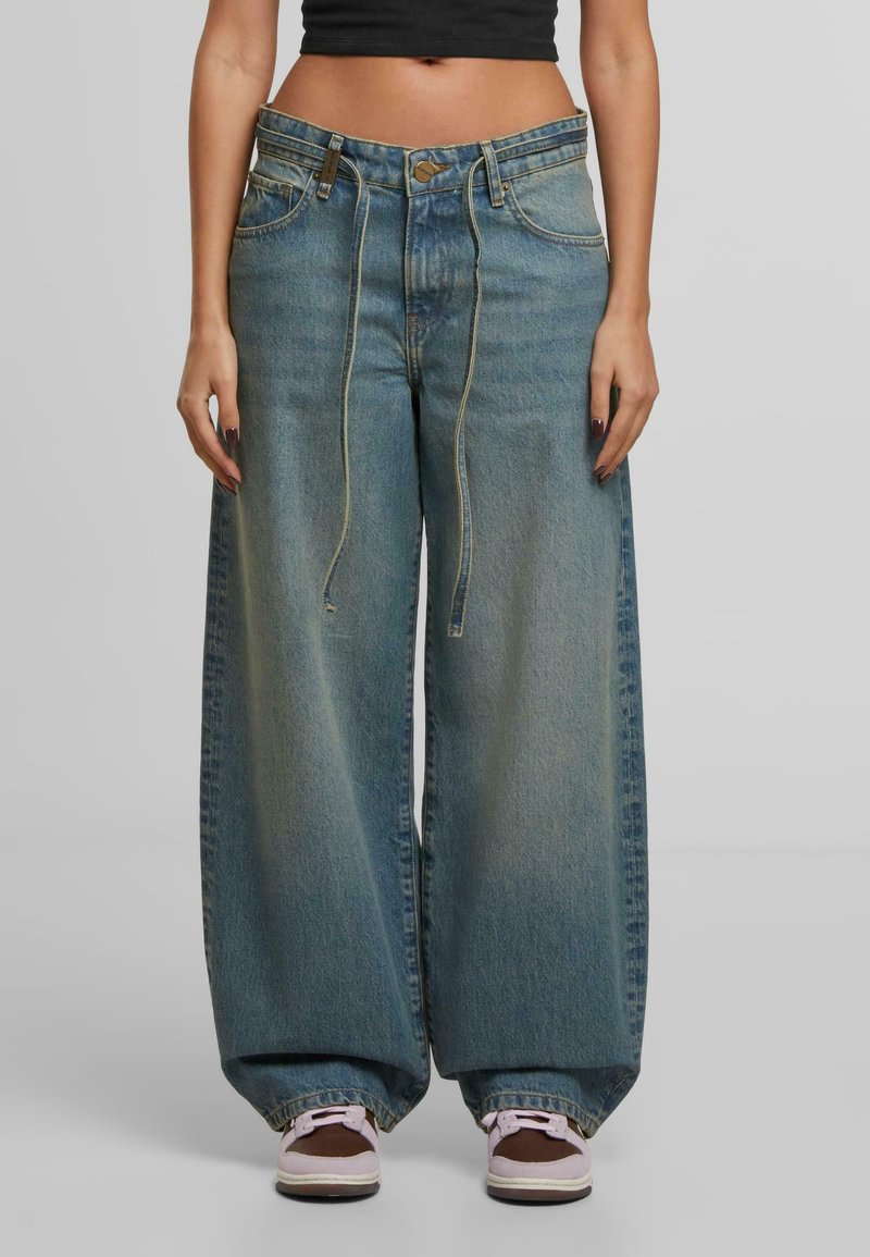 Žena nosi široke plave traperice s elastičnom vezicom na struku i tenisicama, kratki crni top vidljiv na struku.