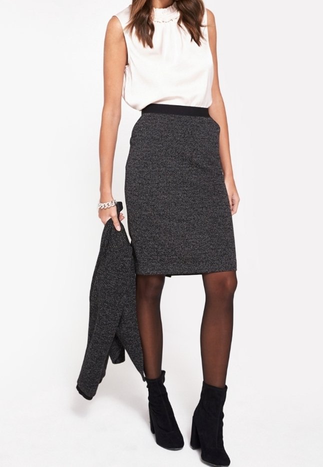 Mujer vestida con una blusa sin mangas blanca, una falda lápiz de tweed gris oscuro, medias negras y botines negros, sosteniendo una chaqueta a juego.