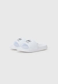 Vita slip-on sandaler i mjukt syntetmaterial, med ett rem med svart Lacoste-logga. Platt sula med texturerad fotbädd.
