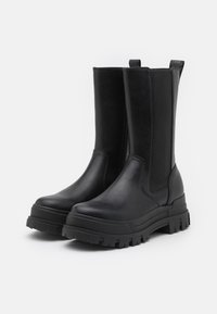 Botas de tobillo de cuero negro con suelas de goma gruesas, paneles laterales elásticos y tiradores, con una textura suave y un diseño minimalista.