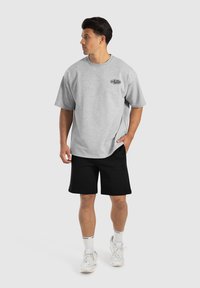 Graues T-Shirt mit kurzen Ärmeln und Logo, kombiniert mit schwarzen Shorts und weißen Turnschuhen. Das Model steht vor einem schlichten Hintergrund.