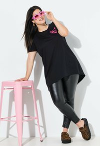 Zwarte oversized t-shirt met roze graphic, zwarte neplederen leggings, luipaardprint klompen en een roze metalen kruk op een witte achtergrond.