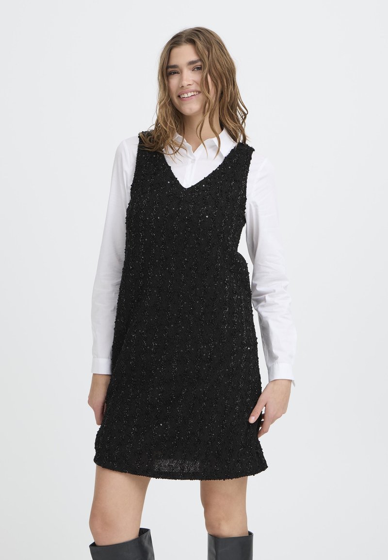 Crna haljina s detaljima od šljokica, V-izrezom i bez rukava, izrađena od teksturiranog materijala s ležernim krojem, preko bijele košulje.