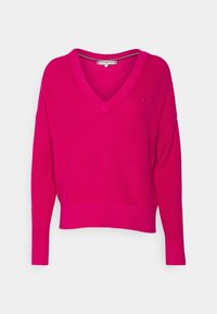 Fuchsia Strickpullover mit V-Ausschnitt, langen Ärmeln und geripptem Saum. Das Modell hat subtile Details an den Seiten und ein Logo-Accent in der Nähe des Ausschnitts.