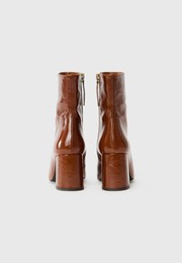 Botas de tobillo de cuero marrón con un acabado brillante, tacones cuadrados y cierre de cremallera en la parte trasera. Textura suave y diseño ajustado.