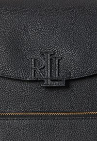 Bolsa de couro preta texturizada com fecho de aba, apresentando um logótipo em relevo e um detalhe em zíper dourado na parte frontal.