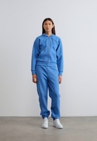 Ανοιχτό μπλε φούτερ με φερμουάρ, συνδυασμένο με ταιριαστά παντελόνια φόρμας. Κατασκευασμένο από μαλακό ύφασμα, διαθέτει κουκούλα με κορδόνι και ελαστικές μανσέτες. Άσπρα αθλητικά παπούτσια.