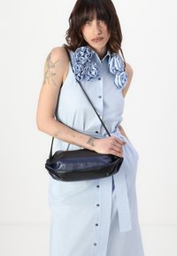 Μαύρη και ναυτική δερμάτινη τσάντα χειρός με ορθογώνιο σχήμα, μαλακή υφή και λεπτό λουρί ώμου, σε στυλ εναντίον ενός ανοιχτού μπλε φορέματος.