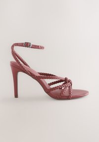 Sandale à talons hauts à bout ouvert en cuir rose poudré. Présente des brides tressées et une boucle de cheville réglable. Texture lisse et silhouette élégante.