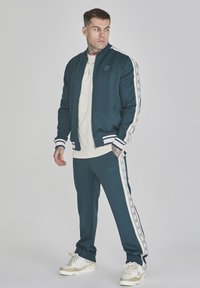 Chándal en color azul verdoso con rayas blancas, chaqueta bomber con cremallera y joggers a juego. Presenta un logo bordado y una tela suave. Camiseta crema debajo.