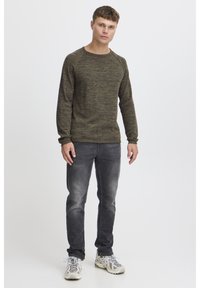 Maglione verde scuro melangiato con polsini e orlo a costine, abbinato a jeans grigi sbiaditi e scarpe da ginnastica chiare su uno sfondo neutro.