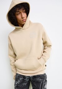 Felpa beige con tasca frontale e logo in rilievo. Presenta un cappuccio con stringhe e un tessuto morbido e texturizzato. Abbinata a pantaloni neri con motivo.