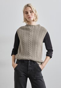 Beige kabelstickad tröja med hög krage, bärs över en svart långärmad topp, stylares med mörka denimbyxor med synliga fickor.