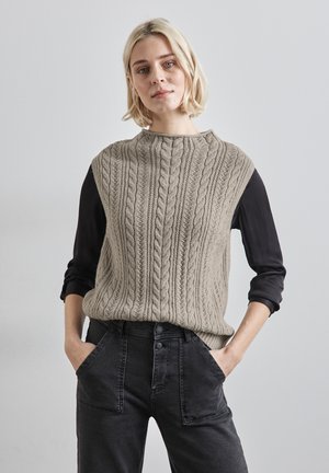 Beżowy sweter w splot kabelkowy z wysokim kołnierzem, noszony na czarnej bluzce z długim rękawem, stylizowany z ciemnoniebieskimi dżinsami z widocznymi kieszeniami.