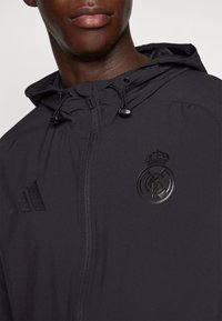Zwarte jas met ritssluiting, voorzien van een capuchon, trekkoorden en reliëflogo's: Adidas op de linkerborst en het Real Madrid-embleem in het midden.