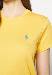 Gul bomullströja med rund halsringning, korta ärmar och en liten blå broderad polo-logga på vänster bröst.