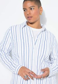 Camicia bianca con righe verticali blu, design con bottoni, maniche lunghe, due tasche frontali e taglio rilassato, indossata sopra un top bianco.