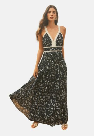 Femeie purtând o rochie maxi neagră cu buline albe, decolteu în V adânc, bretele albe cu margini și sandale albe cu bretele, stând în picioare cu rochia ușor în mișcare.