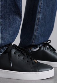 Schwarze Ledersneaker mit weißen Gummisohlen, schwarzen Schnürsenkeln und einem kleinen metallischen Akzent an der Seite. Getragen mit blauen Denim-Jeans.